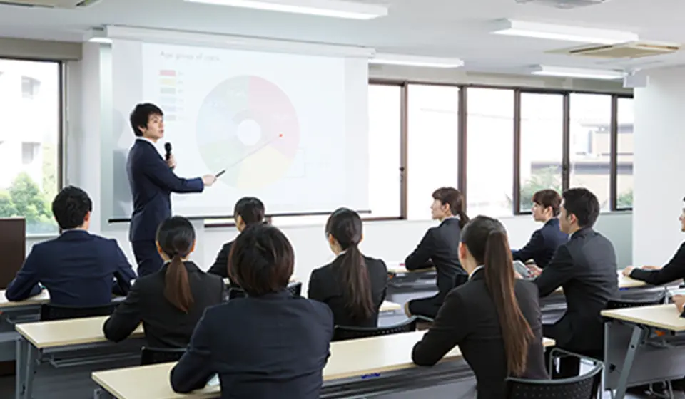 スーツ姿の男性が指し棒を使い、グラフを示しながら研修を行っている様子。